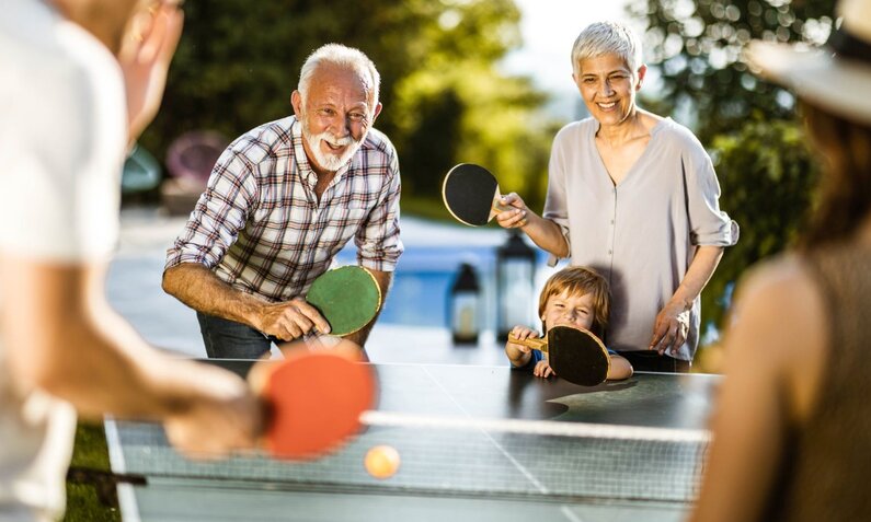 Freude beim Tischtennisspiel im Freien: Ein älteres Paar und ein Junge lächeln und halten Schläger. | © Getty Images / E+ / skynesher