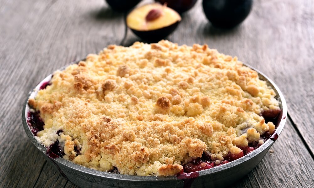 Pflaumencrumble in einer Blechauflaufform, dahinter frische Pflaumen. | © Getty Images / voltan1