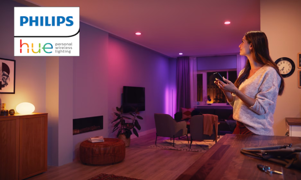 In einem durch Philips Hue farbig beleuchteten Wohnzimmer steht eine Frau mit Smartphone und steuert die Lichter. | © Amazon/Philips