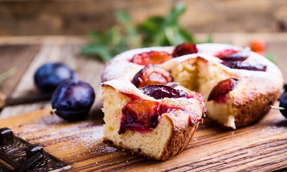 Kleiner angeschnittener luftiger Pflaumenkuchen daneben frische Pflaumen. | © Getty Images / istetiana
