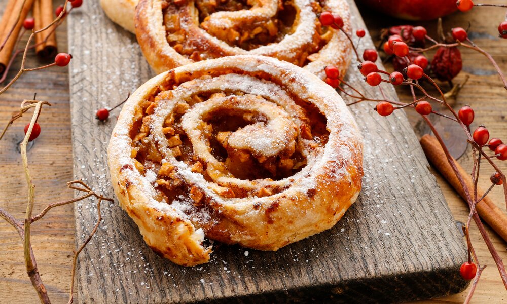 Drei Apfelschnecken auf einem rustikalem Holzbrett angerichtet und mit Puderzucker bestäubt, in herbstlicher Atmosphäre. | © Adobe Stock / agneskantaruk
