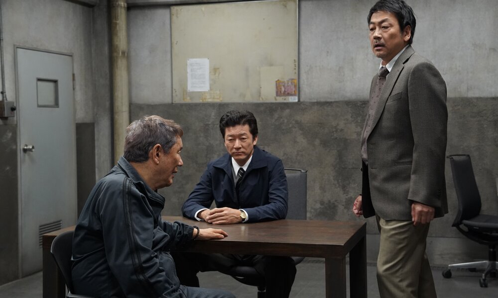 Takeshi Kitano und zwei Co-Stars sitzen und stehen an einem Tisch in einem Verhörraum in einer Szene aus dem Prime-Video-Film "Broken Rage". | © Prime Video/Amazon