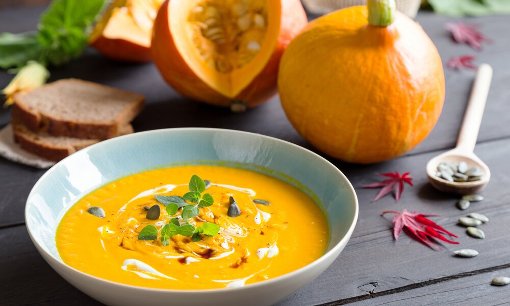 Eine Schüssel voller Kürbissuppe mit Kernen und Kräutern garniert, im Hintergrund ganze Kürbisse. | © Adobe Stock / juefraphoto