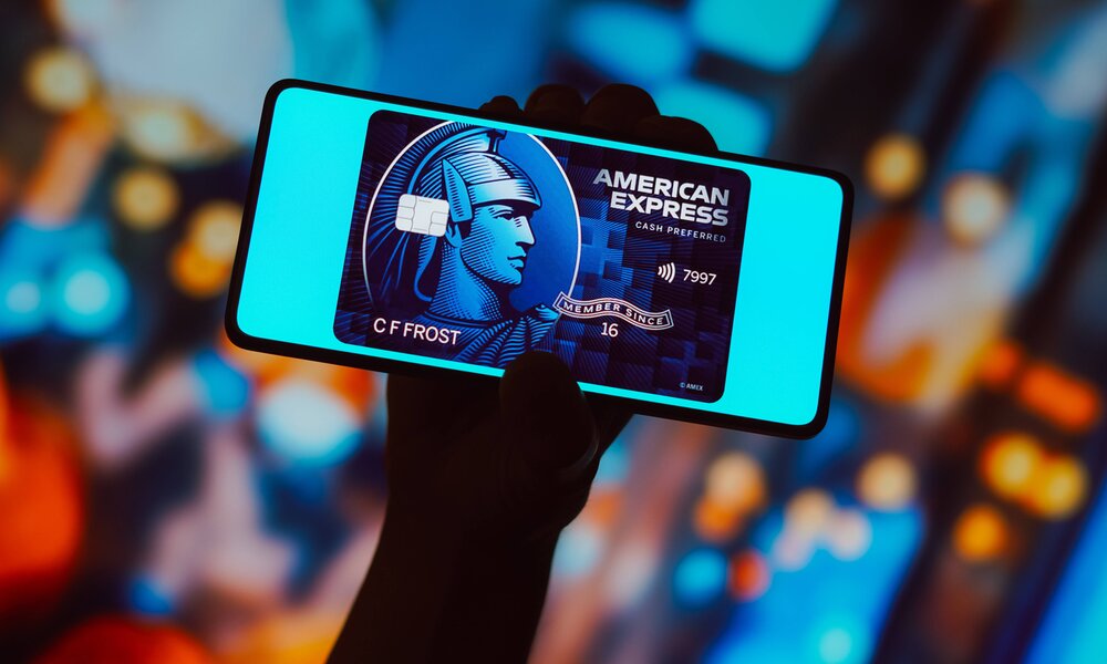 Hand hält Smartphone mit digitaler American Express Blue Card vor verschwommenem, buntem Hintergrund. | © IMAGO/Zoonar