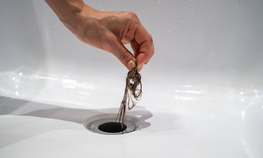 Eine Hand zieht einen nassen, verfilzten Haarknäuel aus einem Abfluss in einem weißen Waschbecken. | © Adobe Stock/ DimaBerlin