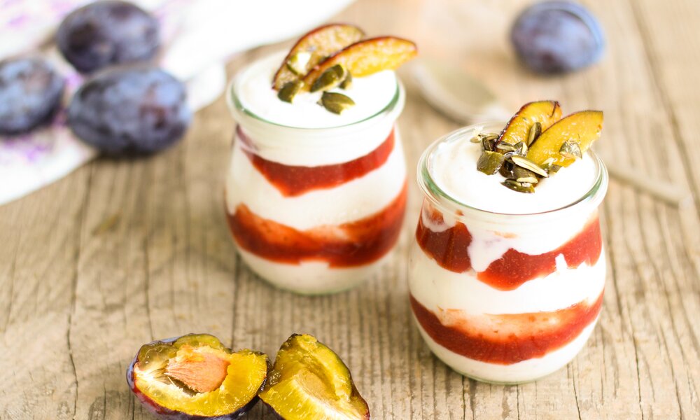 Zwei Pflaumen-Schichtdessert mit frischen Pflaumenkompott und daneben liegen weitere frische Pflaumen. | © Adobe Stock / Bernadette