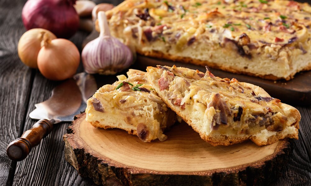 Angeschnittener Zwiebelkuchen auf einem Brett, davor zwei Stücke auf einem Holzbrett. | © Adobe Stock / O.B.