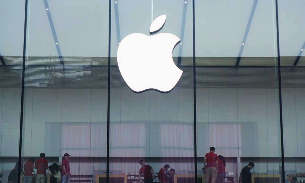 Ein großes weißes Apple-Logo hängt an einer Glasfassade, dahinter Mitarbeiter in roten Shirts in einem Apple-Store. | © Getty Images/ NurPhoto