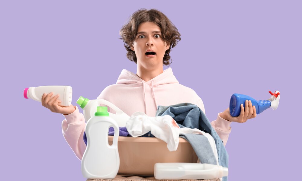 Ein junger Mann mit braunen Locken und erstaunten Gesicht hält zwei Reinigungsmittel in den Händen, vor ihm ein Korb mit Wäsche. | © Adobe Stock/ Pixel-Shot