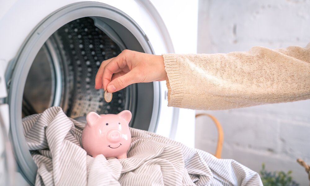 Nahaufnahme einer Hand, die ein Sparschwein in einer Waschmaschine mit einer Münze befüllt, umgeben von gestreiftem Stoff. | © Adobe Stock/ LiettaOraz
