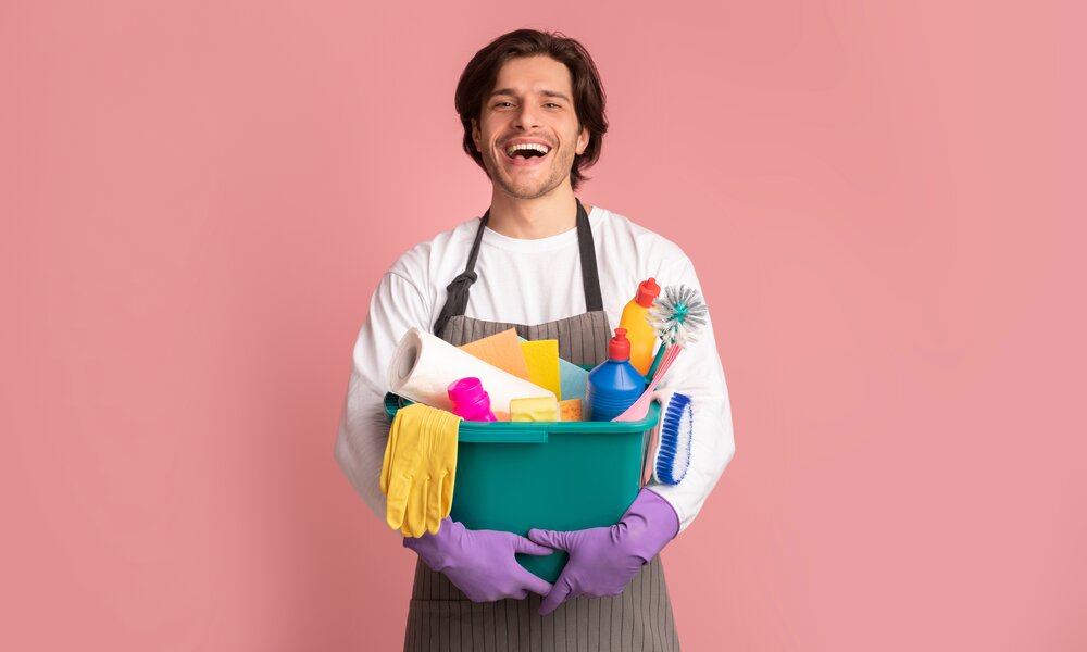 Lachender Mann mit braunem Haar in Schürze und Reinigungshandschuhen hält Putzutensilien in einem grünen Eimer. | © Adobe Stock/ Prostock-studio