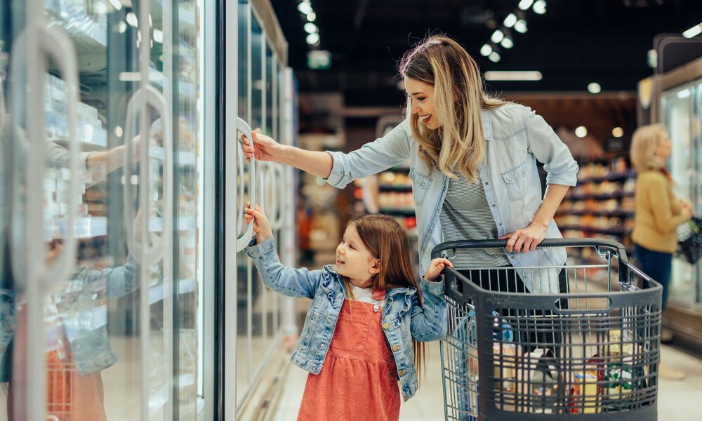 Eine blonde Frau im Jeanshemd öffnet mit einem lächelnden kleinen Mädchen in rotem Kleid die Tiefkühltruhe im Supermarkt. | © Adobe Stock/ bernardbodo