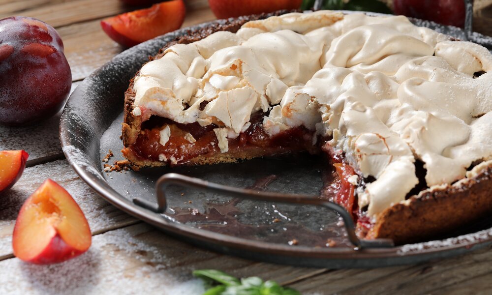 Angeschnittener Pflaumenkuchen mit Baiserhaube in einer Kuchenform, daneben frische Pflaumen. | © Adobe Stock / Leon