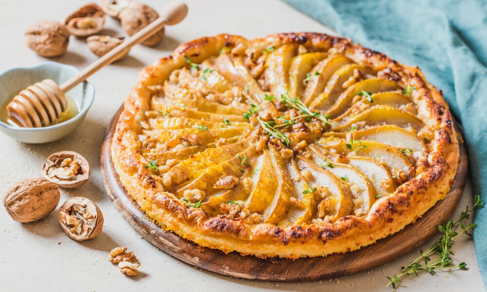 Birnenkuchen mit sauber drapierten Birnenscheiben, darüber glasierte Walnüsse und daneben steht eine kleine Schale mit Honig. | © Adobe Stock / travelbook
