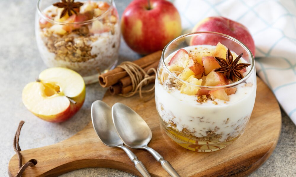 Ein Schichtdessert im Glas, mit weißer Creme und einem Bratapfeltopping, daneben frische Äpfel, Zimt und Sternanis. | © Getty Images / elena_hramowa