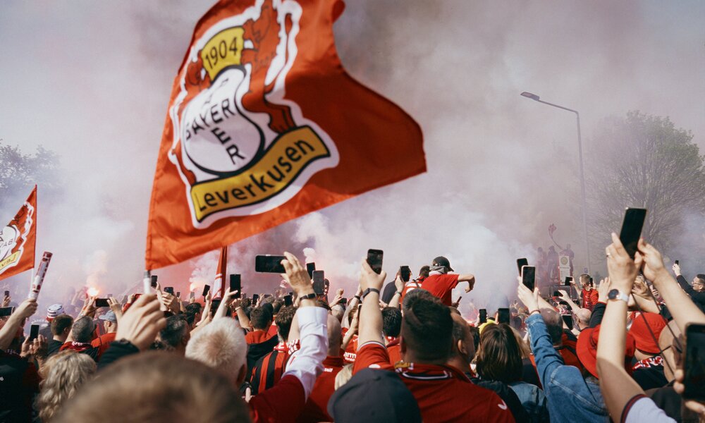 Eine riesege Leverkusenfahne umringt von Fans, die ihre Smartphones hochhalten in einer Szene aus der Prime-Video-Dokuserie "Bayer Leverkusen – A Dream Comes True" | © Amazon Prime Video