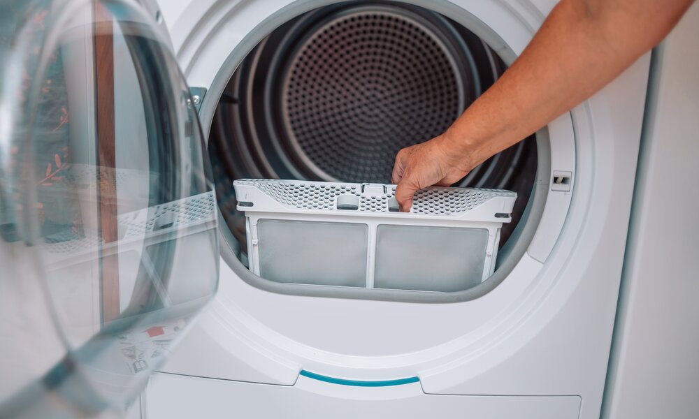 Eine Hand entfernt den Flusenfilter aus der geöffneten Tür eines modernen Wäschetrockners in einem hellen Raum. | © Adobe Stock/ Shi