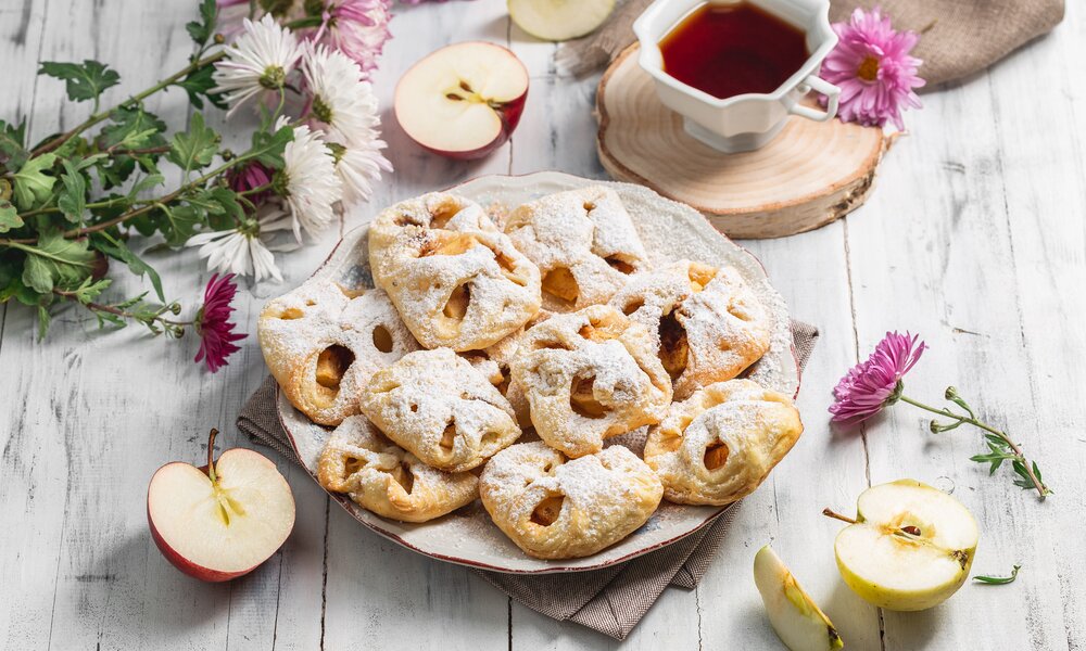 Mehrere süße Blätterteig-Brötchen mit Apfelfüllung auf einem Teller angerichtet, daneben frische Blumen. | © Getty Images / 	AnSyvanych
