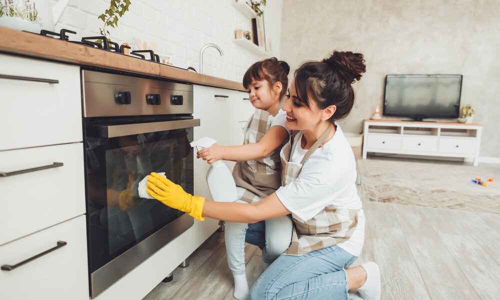 Eine junge Mutter und ihre kleine Tochter reinigen lachend gemeinsam den Backofen in einer modernen Küche. | © Adobe Stock/ inna717