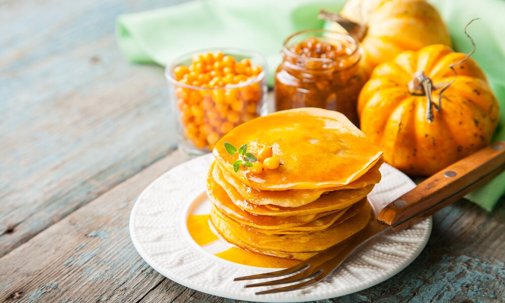 Gestapelte Kürbis-Pfannkuchen auf einem weißen Teller, daneben Gabel und frische Kürbisse. | © Adobe Stock / murziknata
