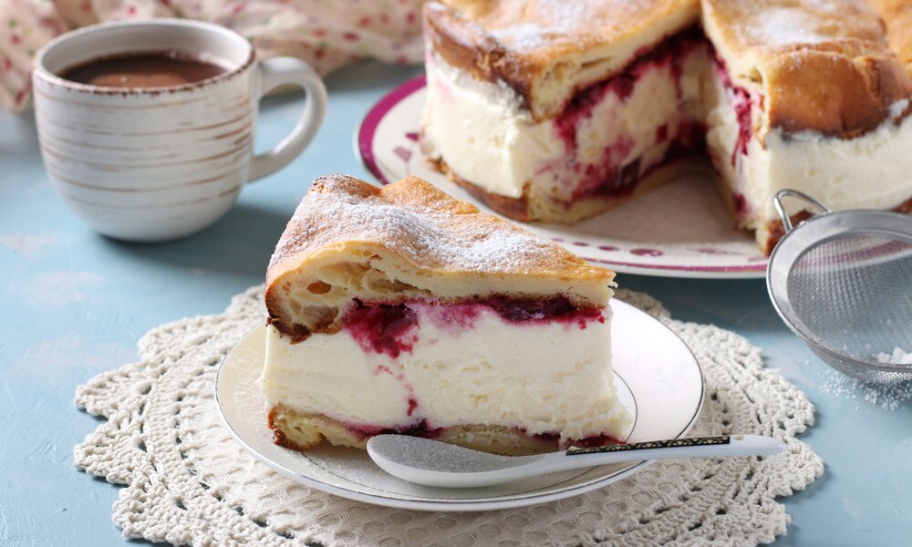 Stück Joghurtkuchen mit Kirschen auf einem Teller, dahinter der angeschnittene, ganze Kuchen. | © Adobe Stock / Anzhela