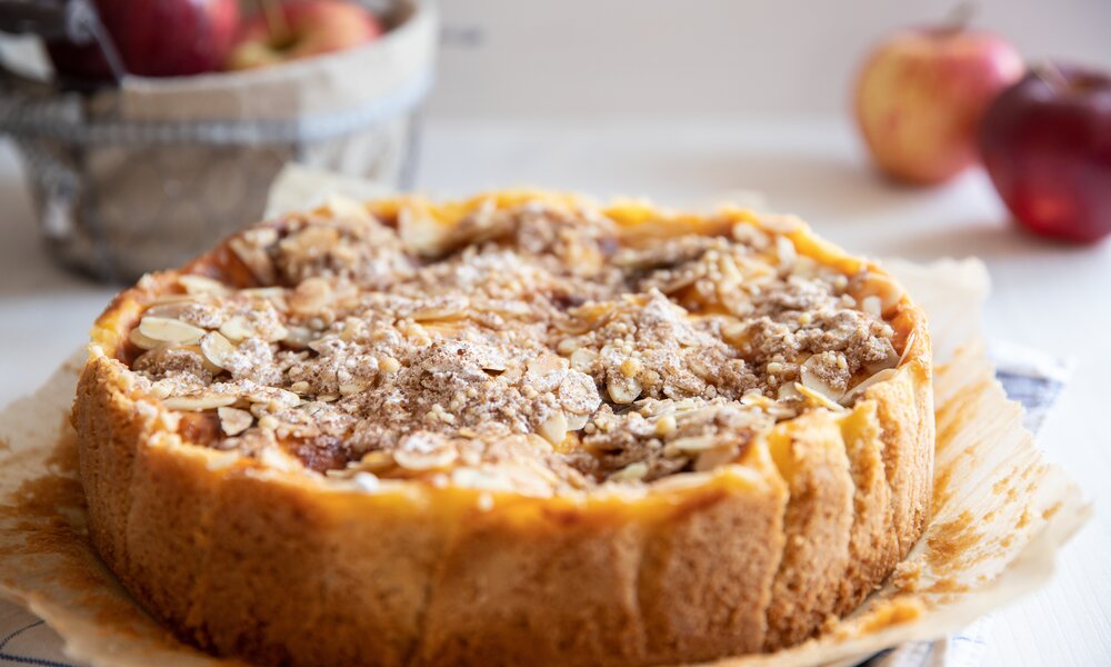 Apfelkuchen auf einem Kuchengitter, darauf Backpapier und im Hintergrund unscharf Äpfel. | © Adobe Stock / Kerstin