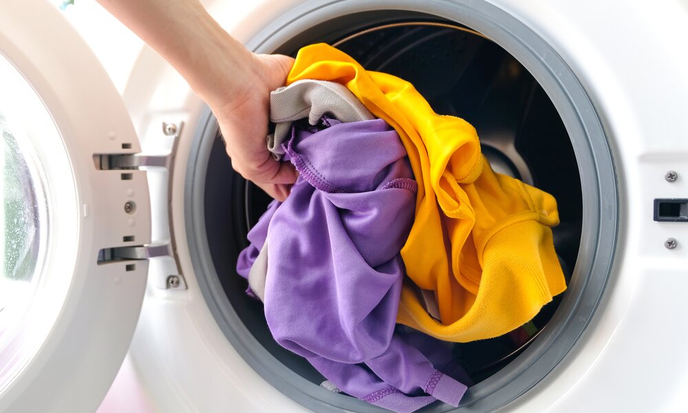 Eine Hand legt bunte Wäsche in Gelb, Lila und Grau in die geöffnete Trommel eines Wäschetrockners | © Adobe Stock/ Photo Sesaon
