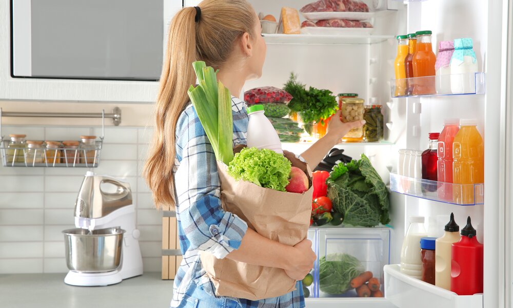 Junge Frau mit blonden Haaren und karierter Bluse räumt Lebensmittel aus einem Papiertüte in einen gut gefüllten Kühlschrank ein. | © Adobe Stock/ New Africa