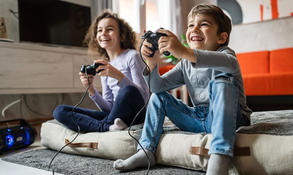Ein Junge und ein Mädchen spielen lachend Videospiele auf dem Boden sitzend, im Hintergrund ein oranges Sofa. | © Miljan Živković/stock.adobe.com