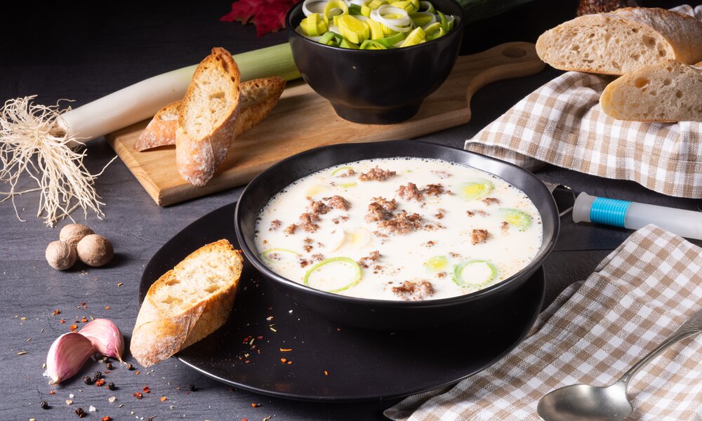 Käse-Lauch-Suppe mit Hackeinlage in schwarzer Suppenschüssel, daneben Baguette, Knoblauch und Lauchzwiebel-Ringe. | © Adobe Stock / Dar1930