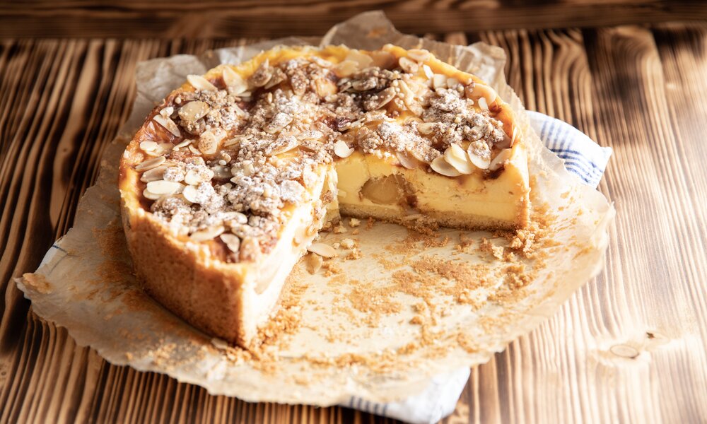 Angeschnittener Apfel-Schmandkuchen auf etwas Backpapier und einem Küchentuch angerichtet, auf einem Holzuntergrund stehend. | © Adobe Stock / Kerstin