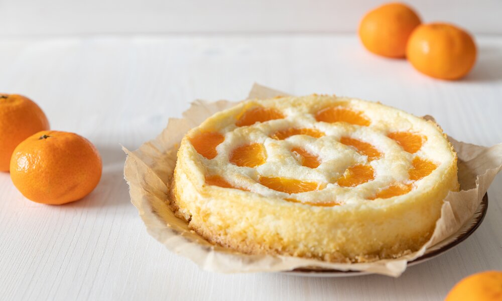 Fluffiger Mandarinen-Schmandkuchen auf einem mit Backpapier belegtem Teller, daneben frische Mandarinen. | © Adobe Stock / Kerstin