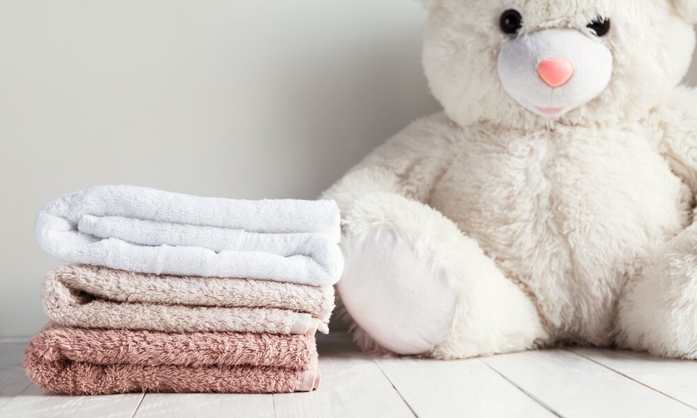 Ein großer cremefarbener Teddybär sitzt neben drei gefalteten Handtüchern in Weiß, Beige und Rosa auf einem weißen Holzboden. | © Adobe Stock/ vladdeep
