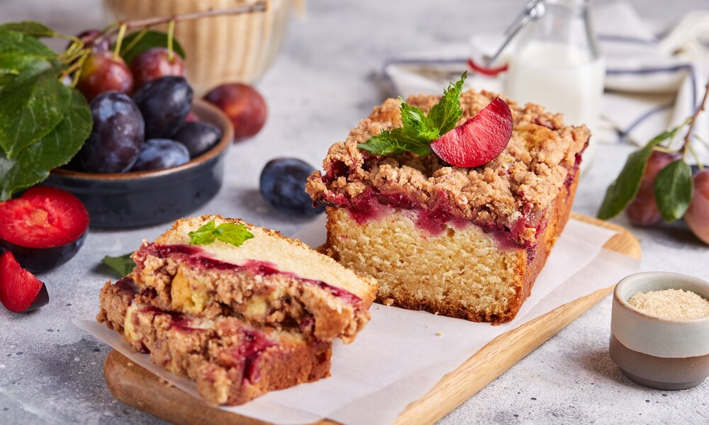 Angeschnittener Kuchen mit Pflaumen in herbstlicher Ambiente, dahinter frische Pflaumen und Milch. | © Adobe Stock / natkinzu