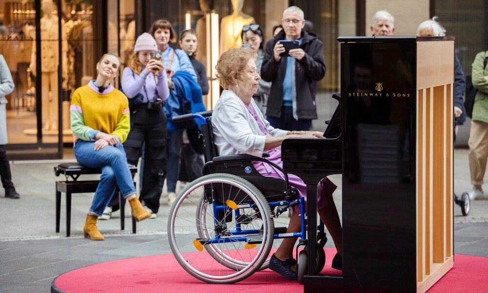 Eine ältere Dame in einem lilafarbenen Kleid spielt im Rollstuhl Klavier, umgeben von einer aufmerksamen Menschenmenge auf einem belebten Platz. | © RTL/Hendrik Lüders