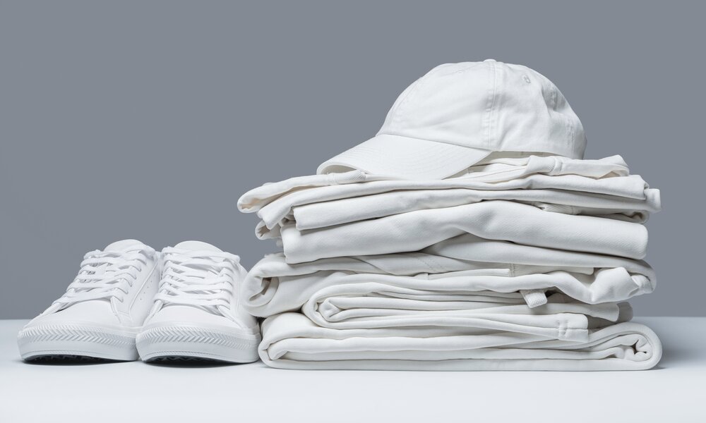 Ein Stapel weißer, gefalteter Kleidung mit weißer Kappe daneben, links stehen weiße Turnschuhe auf einem hellgrauen Hintergrund. | © Adobe Stock/ blackday