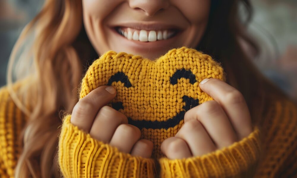 Frau in gelbem Strickpullover hält ein gehäkeltes Smiley-Gesicht und lächelt breit, Nahaufnahme ihres Gesichts. | © Yiss Design/stock.adobe.com