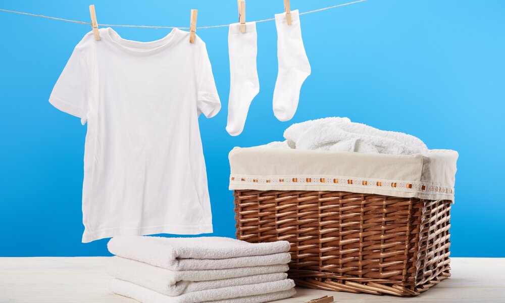 Ein weißes T-Shirt und Socken hängen an einer Wäscheleine vor blauem Hintergrund, daneben ein Korb mit Handtüchern. | © Adobe Stock/ LIGHTFIELD STUDIOS