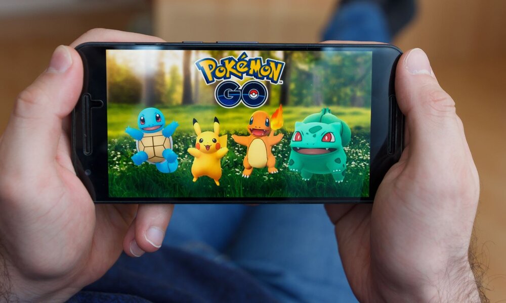 Eine Person hält ein Smartphone mit der App "Pokémon Go" offen, auf dem Bildschirm sind vier Pokémon vor einer Waldszene zu sehen. | © vladim_ka / stock.adobe.com