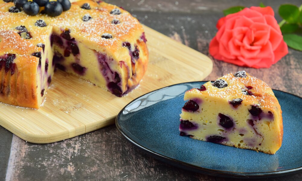 Angeschnittener Blaubeerkuchen auf einem Kuchenteller, davor auf einem Teller ein Stück des Kuchens. | © Adobe Stock / AmalliaEka