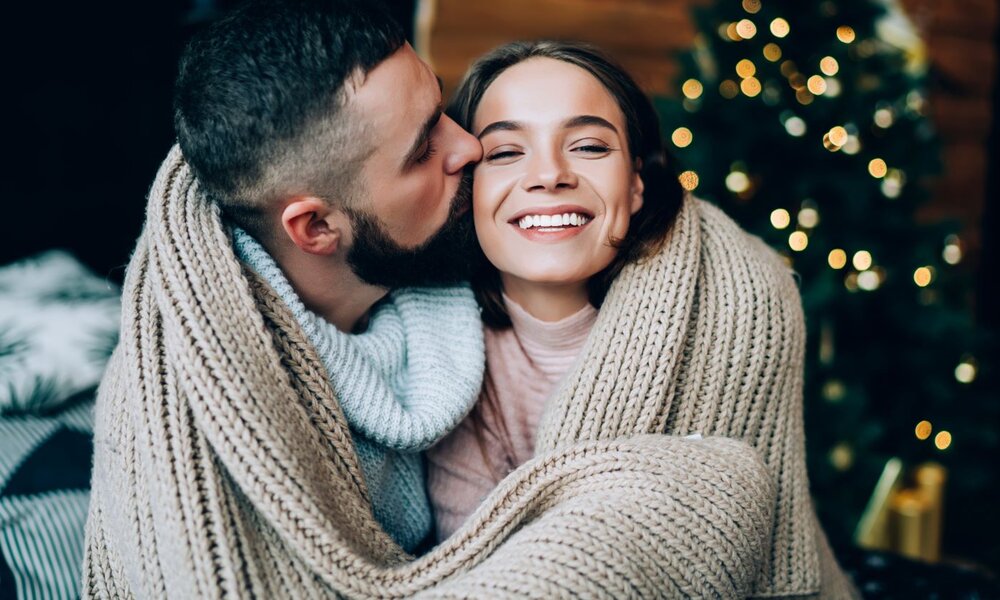 Ein Mann mit Bart küsst eine lachende Frau auf die Wange, beide in warme Decken gehüllt, vor einem geschmückten Weihnachtsbaum. | © GalakticDreamer/ stock.adobe.com