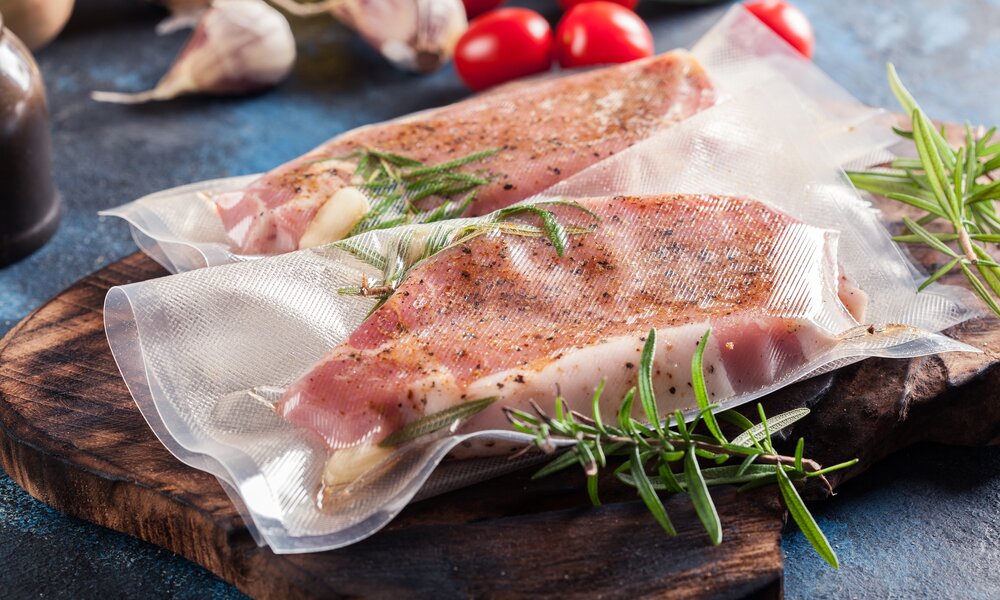 Zwei vakuumierte, gewürzte Schweinesteaks mit Rosmarin und Knoblauch liegen auf einem rustikalen Holzbrett, umgeben von Tomaten. | © Adobe Stock/ Sławomir Fajer