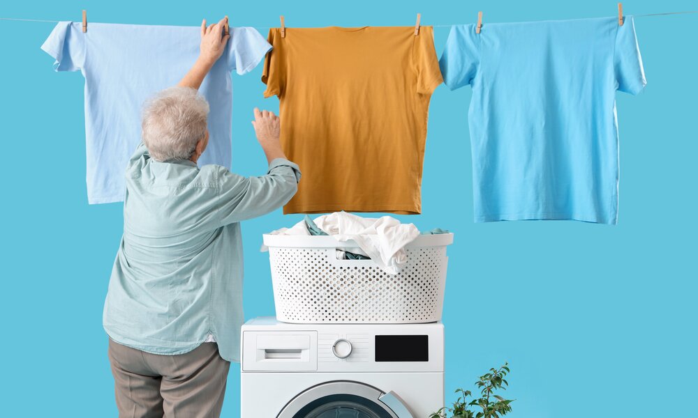 Eine ältere Frau mit grauen Haaren hängt ein blaues und ein senfgelbes T-Shirt an eine Wäscheleine über einer Waschmaschine. | © Adobe Stock/ Pixel-Shot