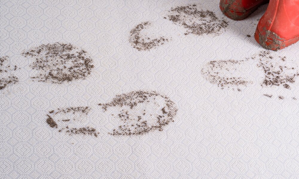 Schlammige Fußabdrücke auf einem weißen Teppich mit geometrischem Muster, daneben stehen rote, schmutzige Gummistiefel. | © Adobe Stock/ HalynaRom