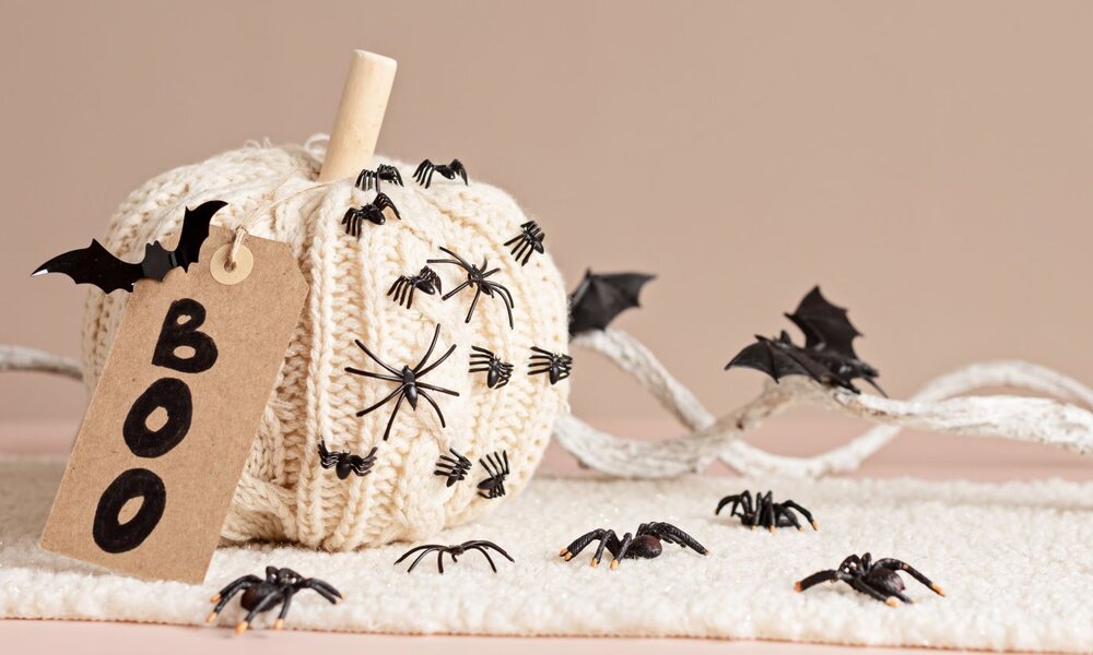 Weißer gestrickter Kürbis, dekoriert mit schwarzen Plastikspinnen und Fledermäusen, daneben ein Etikett mit der Aufschrift "Boo" auf beigem Hintergrund. | © netrun78/stock.adobe.com