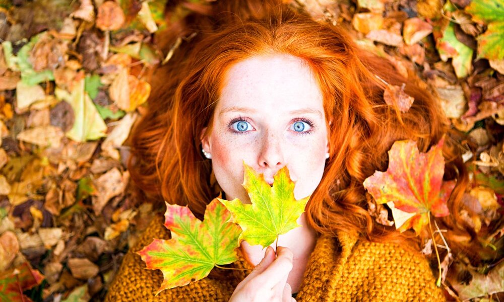 Junge Frau mit roten Haaren liegt lächelnd auf herbstlichem Laub, hält zwei bunte Ahornblätter vor ihr Gesicht und schaut in die Kamera. | © drubig-photo/stock.adobe.com