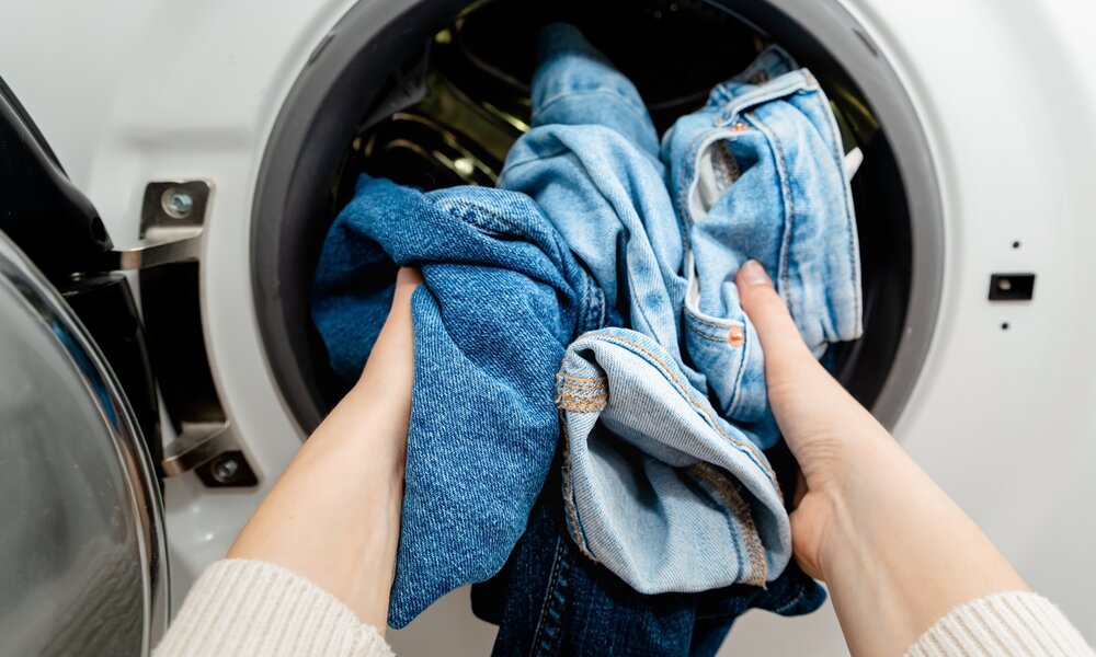 Nahaufnahme von Händen, die hell- und dunkelblaue Jeans in eine Waschmaschine legen. | © Adobe Stock/ Sappheiros