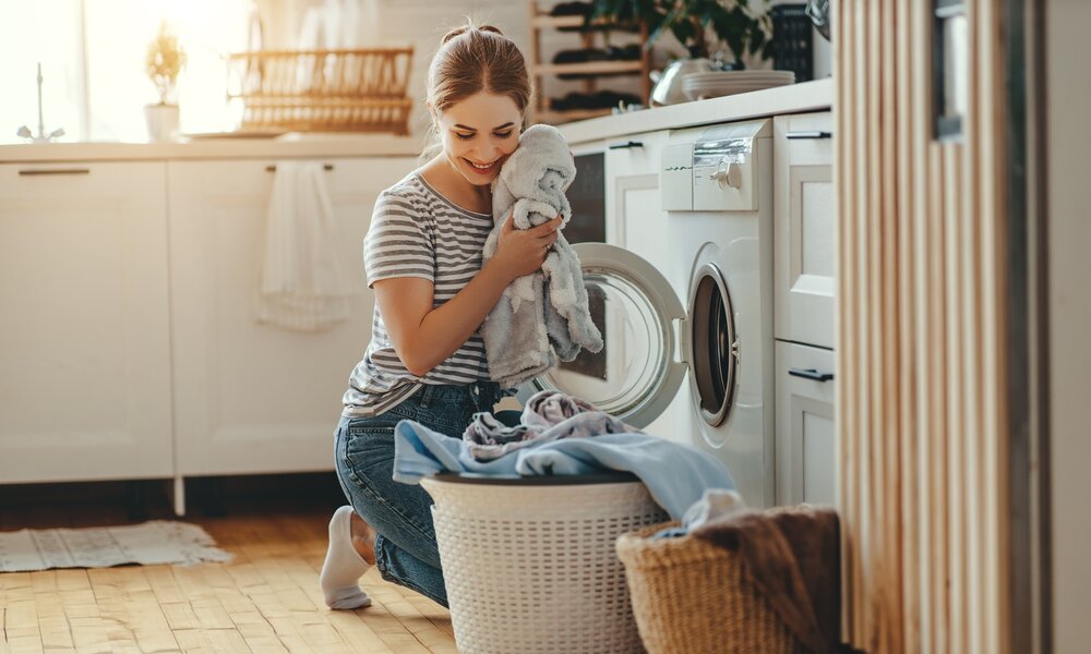 Eine junge Frau in Jeans und gestreiftem T-Shirt kniet lächelnd vor der Waschmaschine und drückt frische Wäsche an ihr Gesicht. | © Adobe Stock/ JenkoAtaman