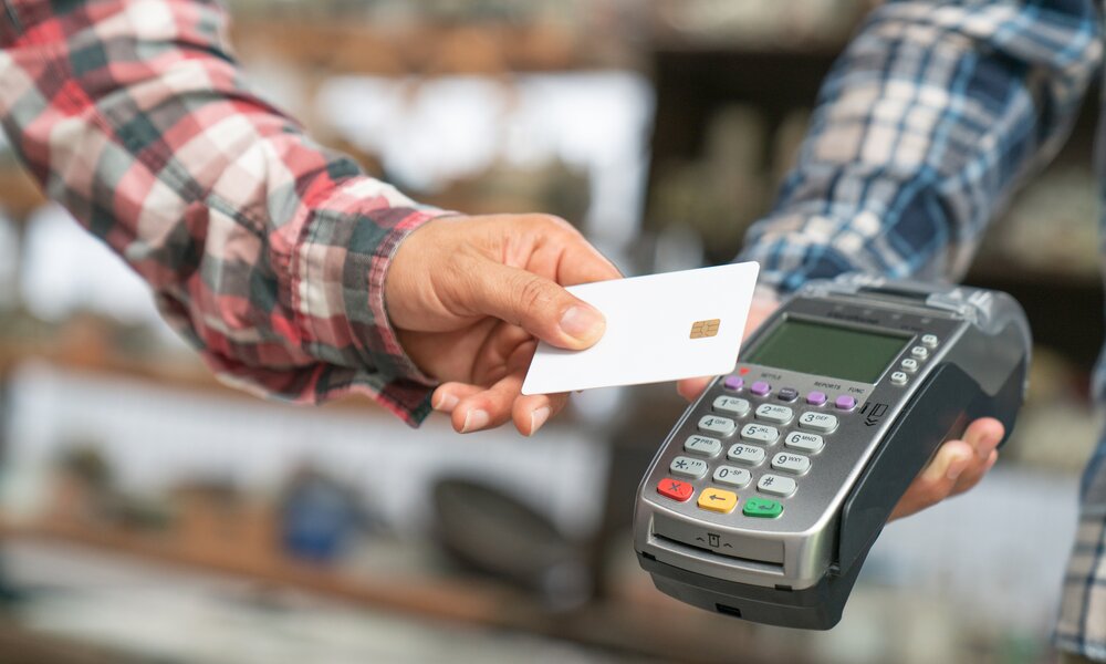 Nahaufnahme einer Person in kariertem Hemd, die eine Kreditkarte an ein EC-Terminal zum kontaktlosen Zahlen hält. | © Getty Images/Witthaya Prasongsin