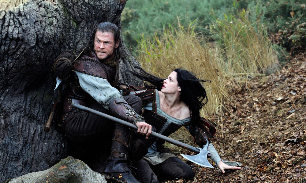 Chris Hemsworth und Kristen Stewart verstecken sich in mittelalterlicher Kleidung hinter einem Baumstamm in einer Szene aus dem Kinofilm "Snow White and the Huntsman" | © Imago Images/Alex Bailey/Universal Pictures/Everett Collection